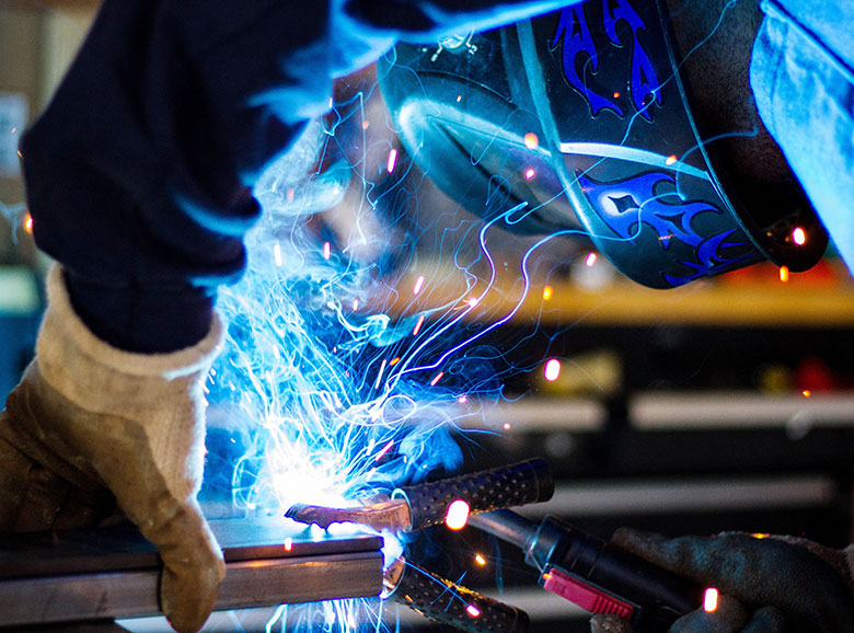 Schweißer mit Handschuhen und Helm verschweißt Metall, leuchtende blaue Funken sprühen in Werkstattumgebung.