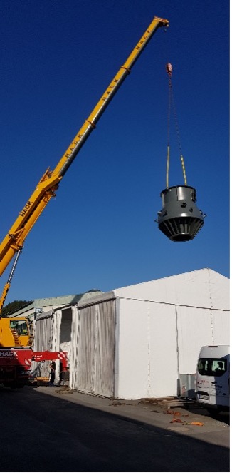 Ein gelber Kran hebt ein großes Filtertechnik-Bauteil über ein weißes Gebäude mit geparktem Lkw bei blauem Himmel.