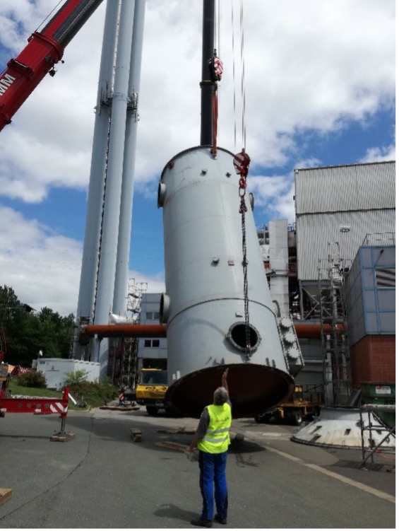 Arbeiter mit gelber Weste weist auf dem Industriegelände einen von einem Kran angehobenen Filtertechnik-Tank ein.