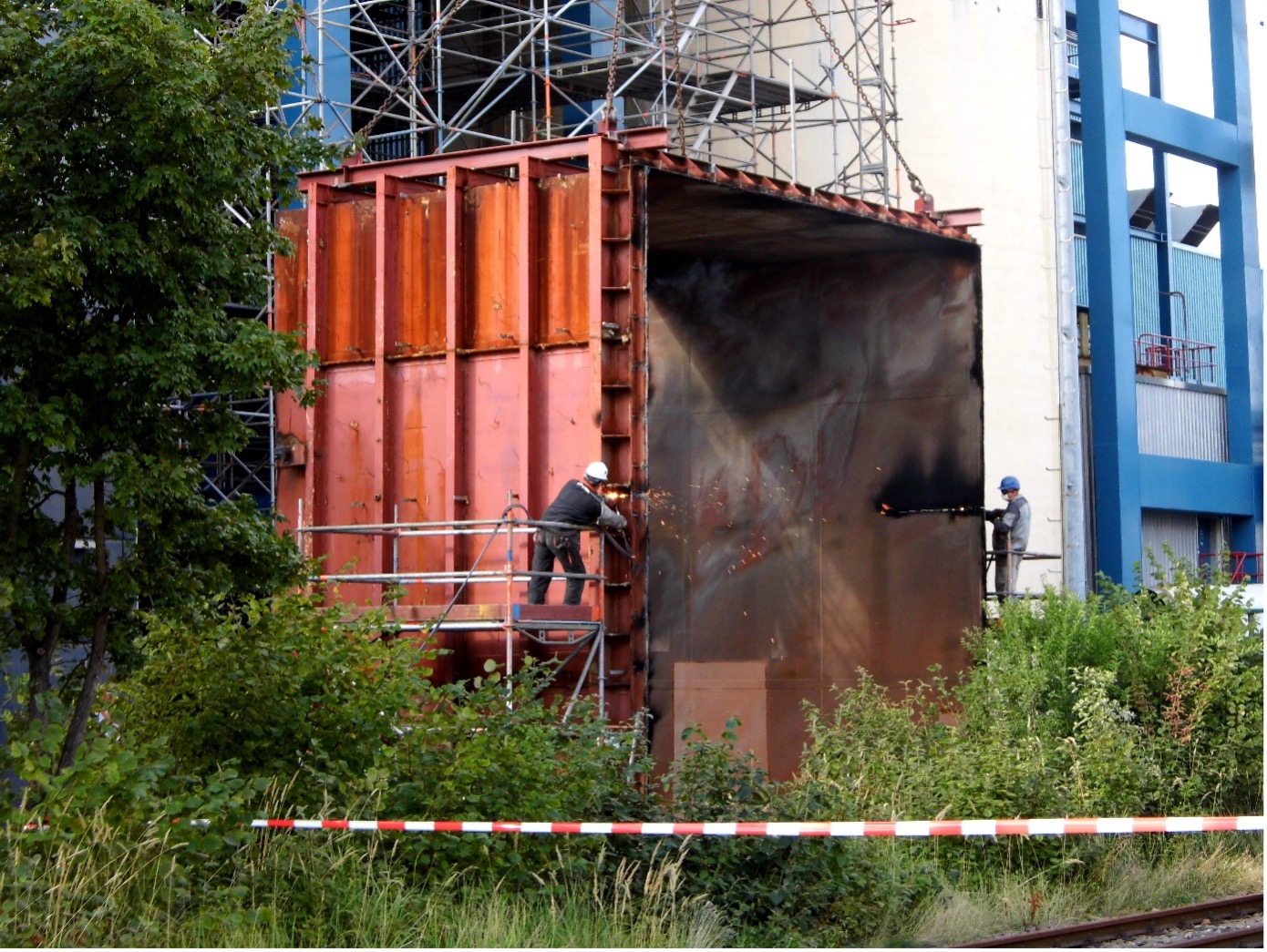 Zwei Arbeiter in Schutzkleidung schneiden eine rostige Metallkonstruktion bei Gerüst, Bepflanzung und Warnband.