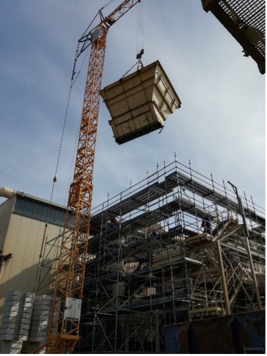 Gelber Baukran hebt großen Filtertechnik-Container neben eingerüstetem Gebäude bei klarem Himmel auf.