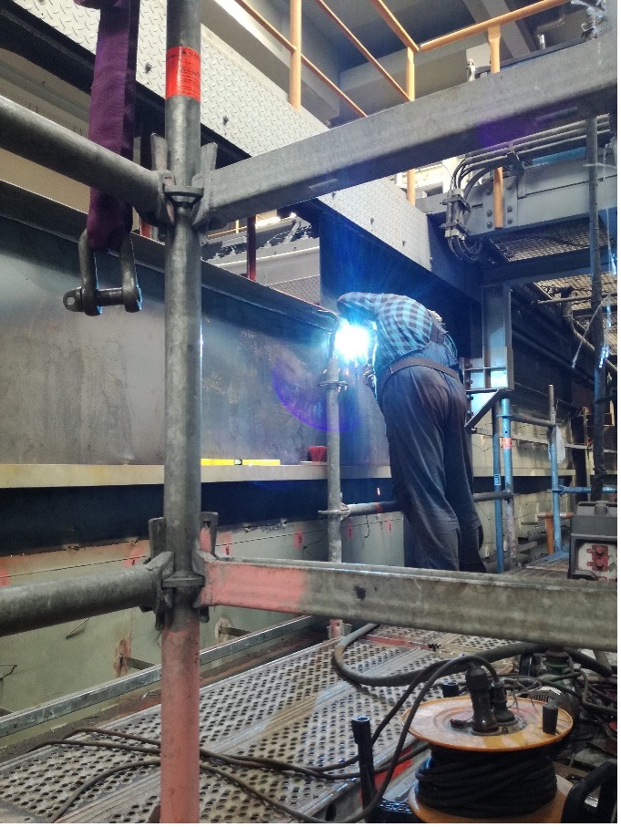 Person in Schutzkleidung schweißt Metallträger mit Filtertechnik auf Baustelle, umgeben von Gerüsten.